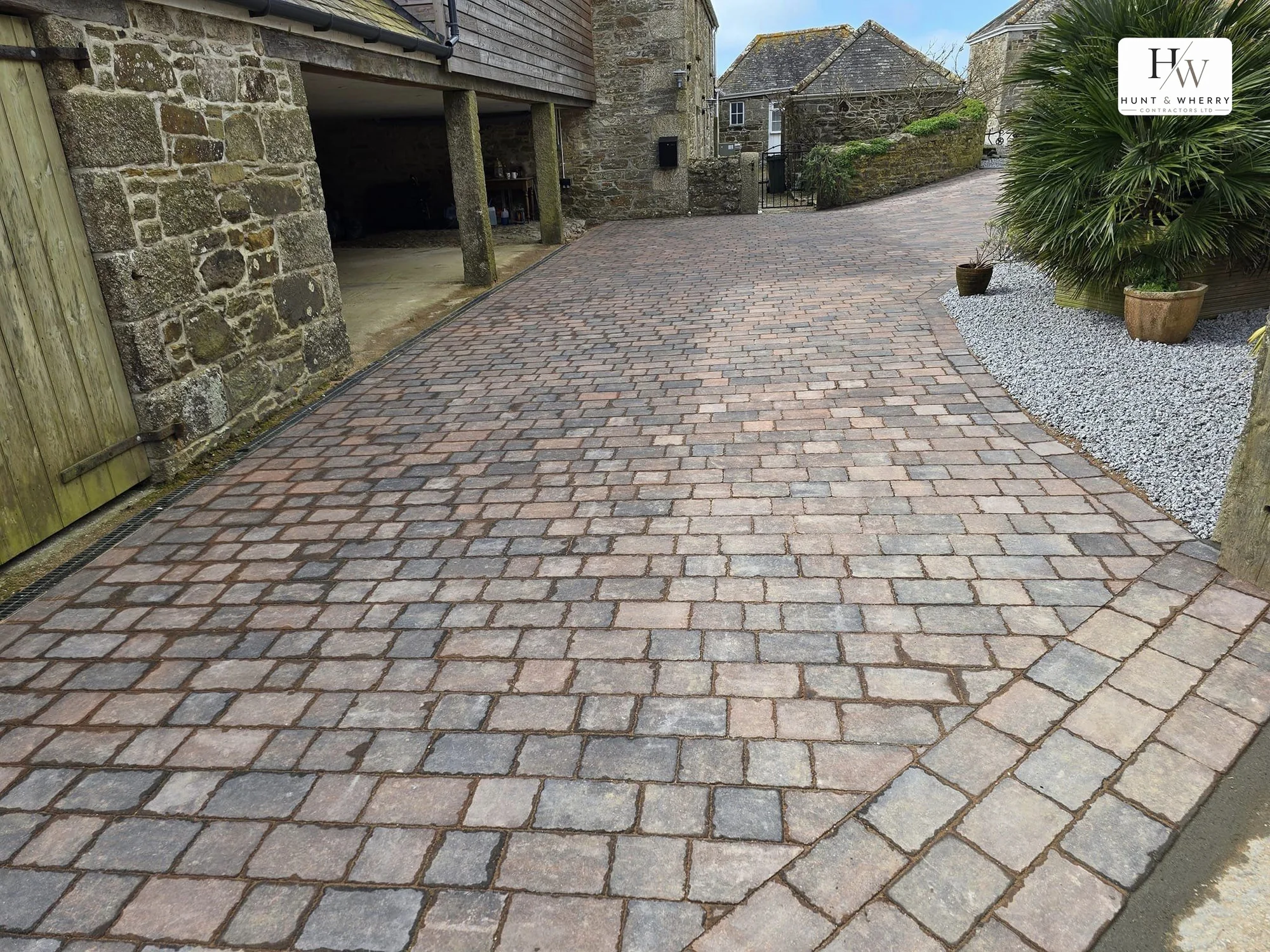 Tumbled cobble sett driveway laid through a Cornish granite barn complex, with a linear drainage channel along the barn wall