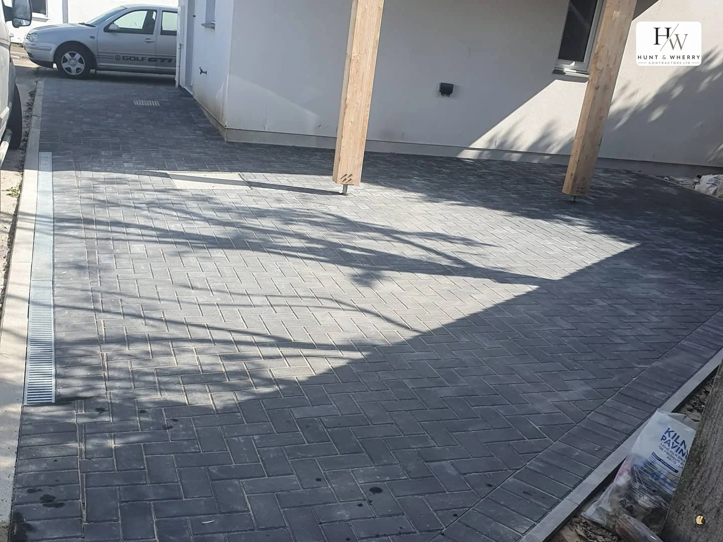 Charcoal and grey herringbone block paving under a timber carport, with a galvanised linear drainage channel