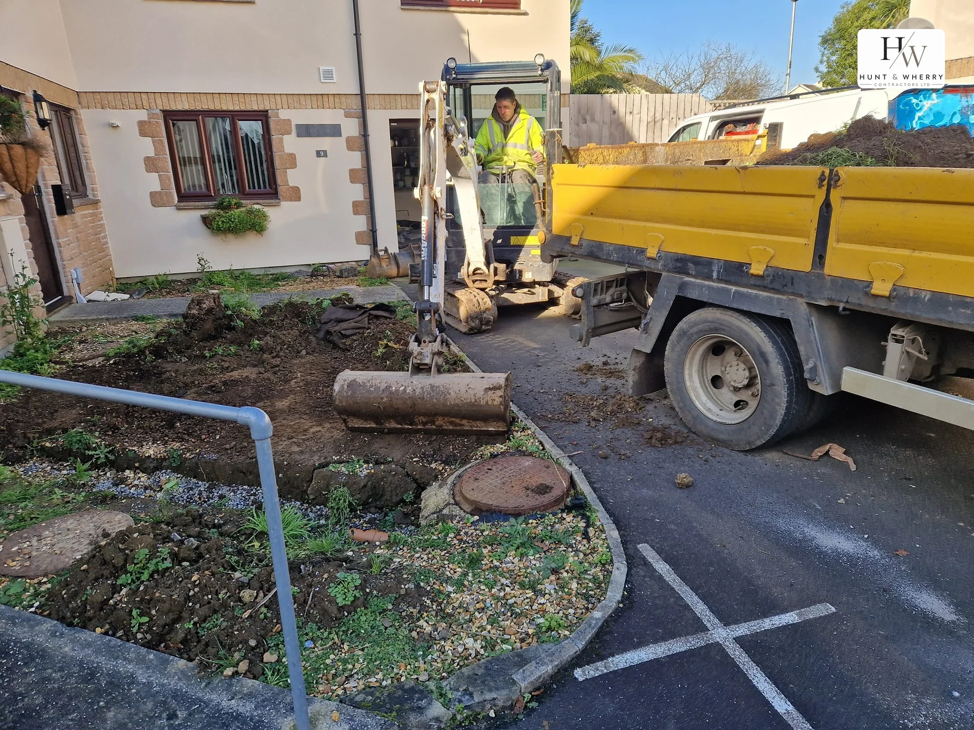 Exposed manhole chamber during drainage work, with a mini excavator and a loaded tipper truck at the kerbside