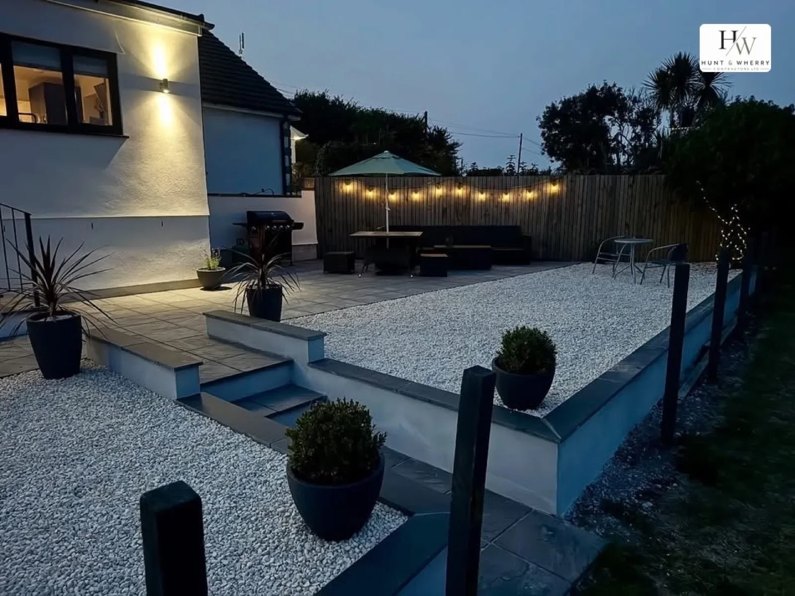 Landscaped garden at dusk with raised patio, white gravel and festoon lighting