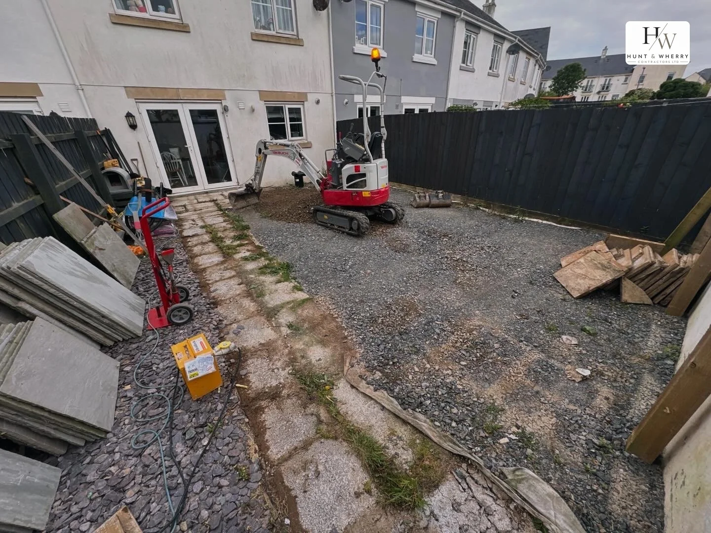 Mini digger working in a rear garden on a compacted stone sub-base, with membrane part laid and paving slabs stacked ready