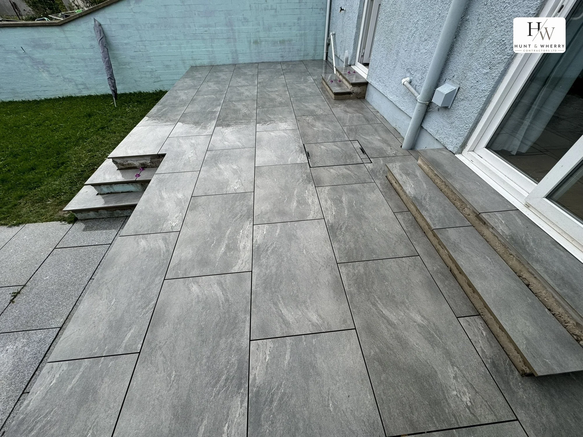 Large format grey porcelain paving laid to a rear terrace, with steps down to the lawn and recessed manhole covers