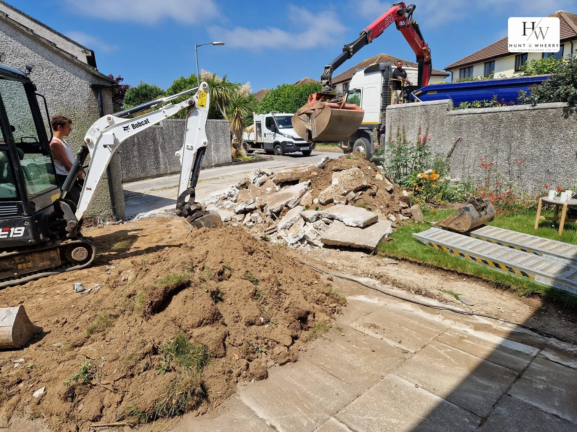 Old concrete driveway being broken out by a mini excavator while a grab lorry loads the spoil from the highway